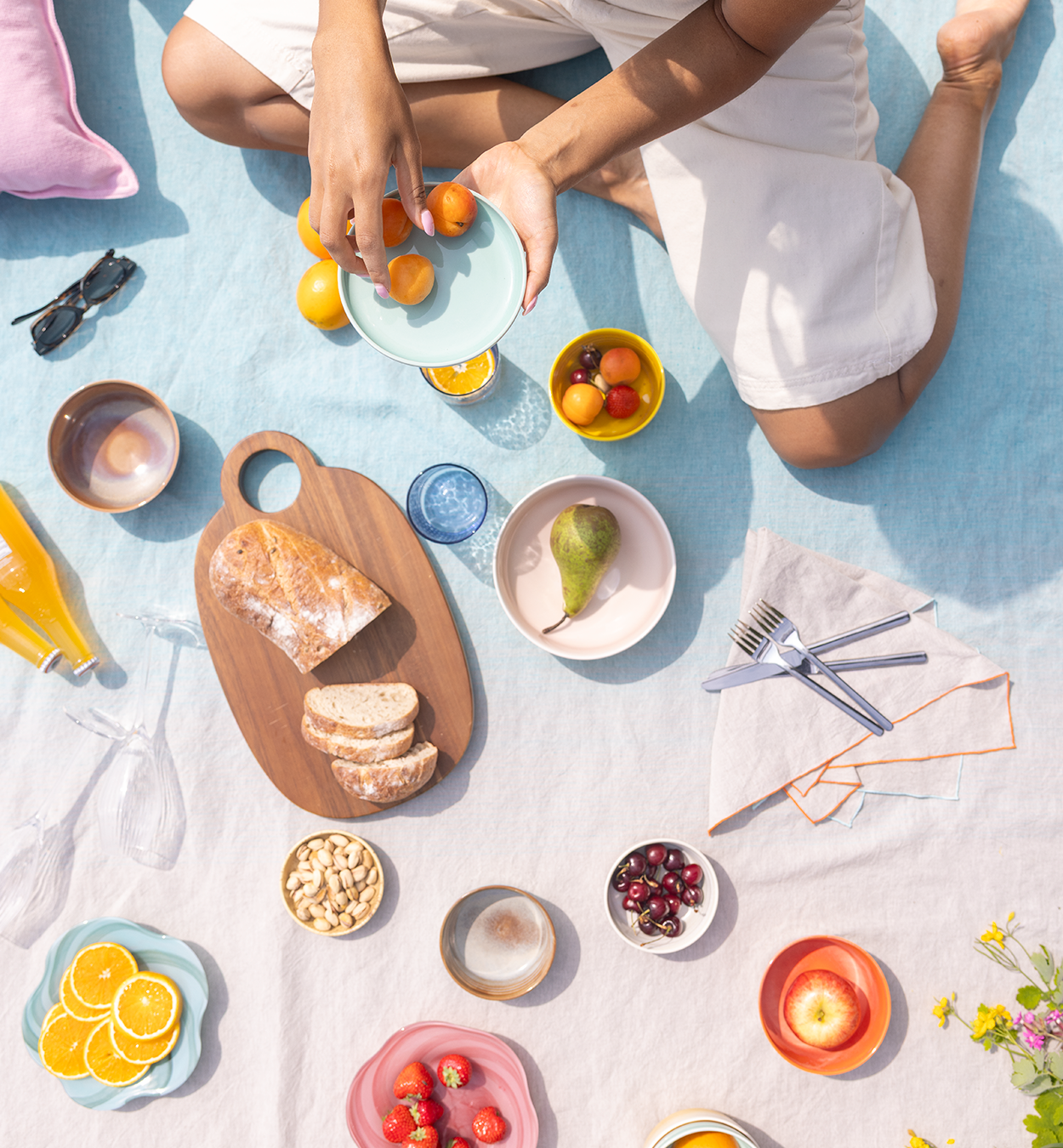 Colorful and playful picnic setting on a light blue linen tablecloth. Woman holding a light turquoise Tuntu plate with yellow plums on it.