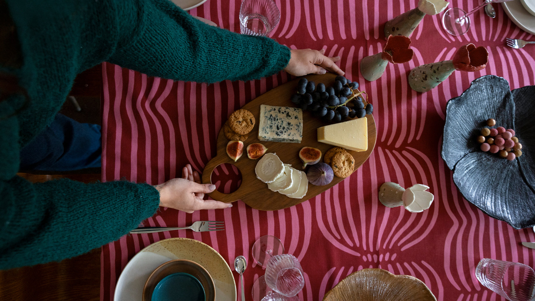 Nainen laskee pöytään Lautta-leikkuulaudan päälle kootun juustoasetelman. Pöydässä punainen Vilja-pöytäliina, Tuntu-astioita ja Anu Pentikin tekemiä Joulutulppaani-keramiikkaveistoksia. Ohjaus joulun kattaus -tuotekategoriaan.